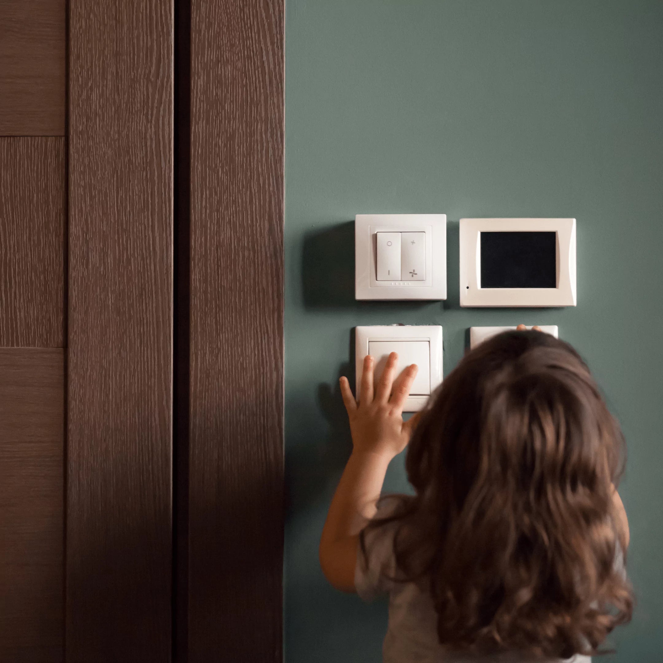 Young child reaching up to press assorted wall switches and controls on a green wall