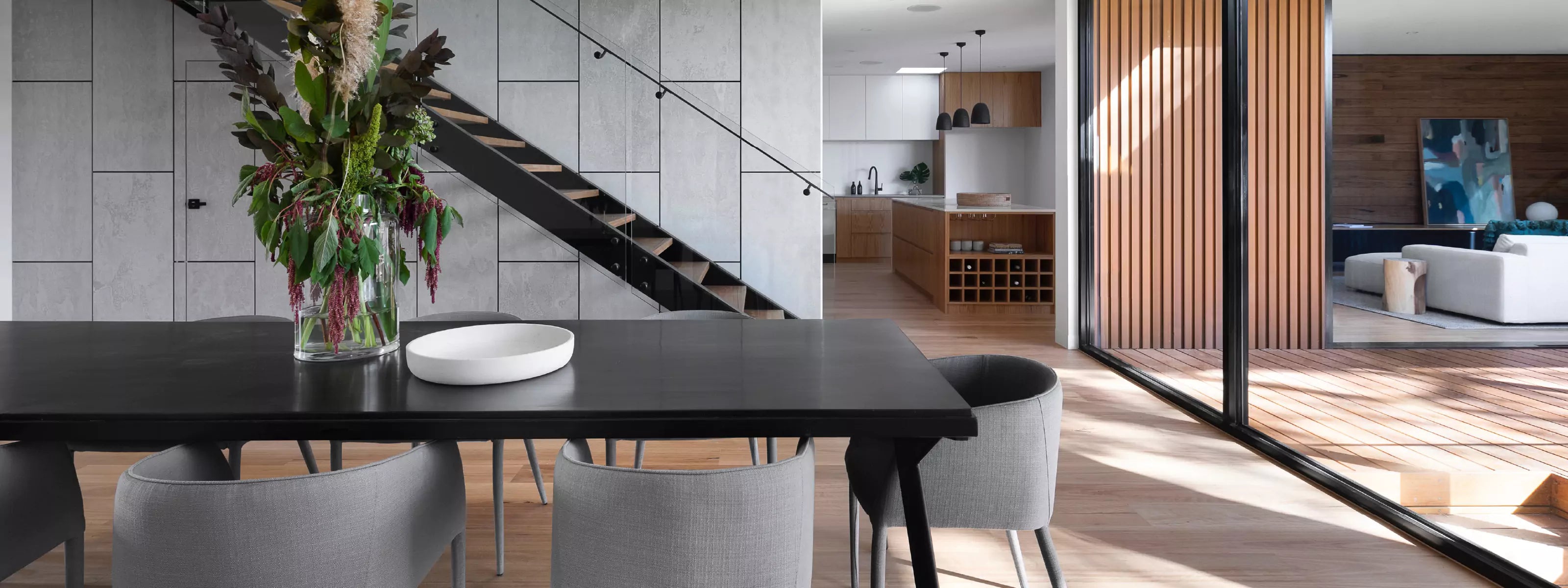 Modern dining space with a minimalist black table, gray seating, and wood accents with an open view to the kitchen and patio
