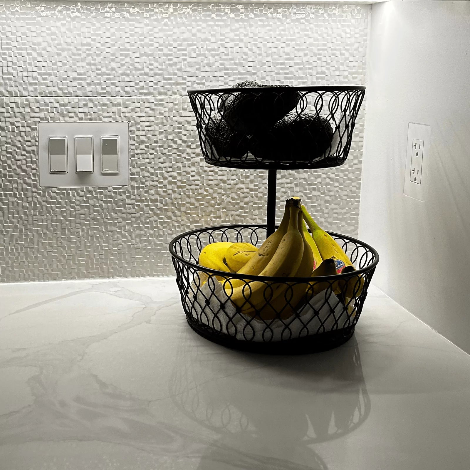 Three light switches CONCEAL flush-mounted on a white tile backsplash behind a black fruit basket on a marble countertop.