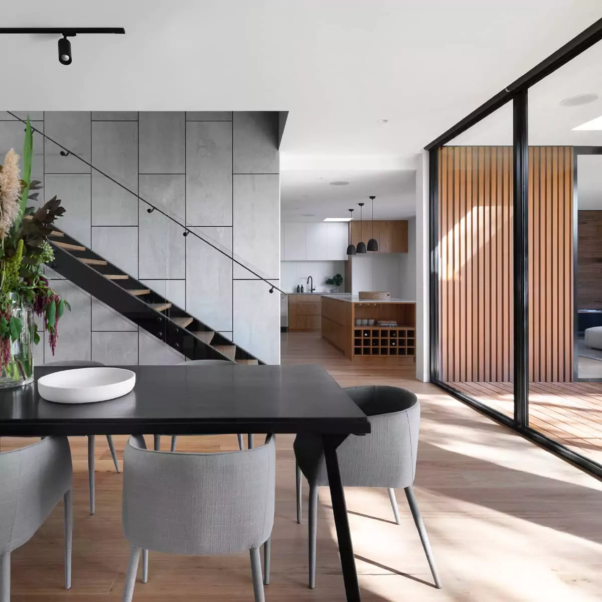 Modern dining space with a minimalist black table, gray seating, and wood accents with an open view to the kitchen and patio