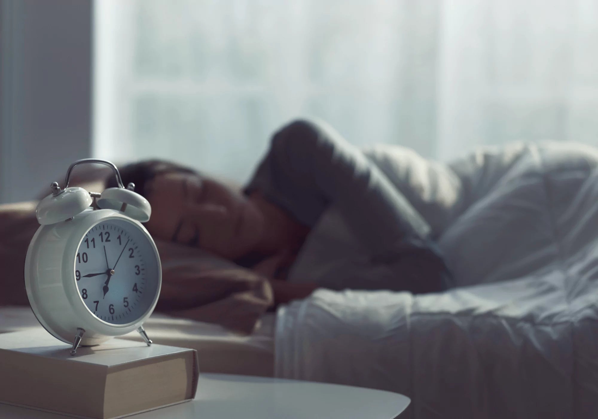 Woman sleeping beside a classic alarm clock, illustrating automated wake-up lighting schedules made possible by ALLY